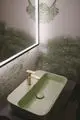 Waschbecken in sanften Grüntönen mit einer goldenen Armatur von Ideal Standard. Die Wand ist mit einem grünen Baumdesign dekoriert.